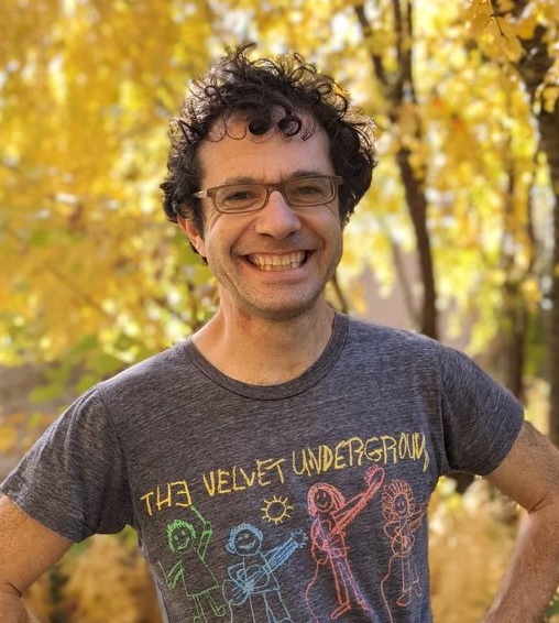 Ben Urwand smiling outdoors in a Velvet Underground T-shirt, standing in front of autumn trees